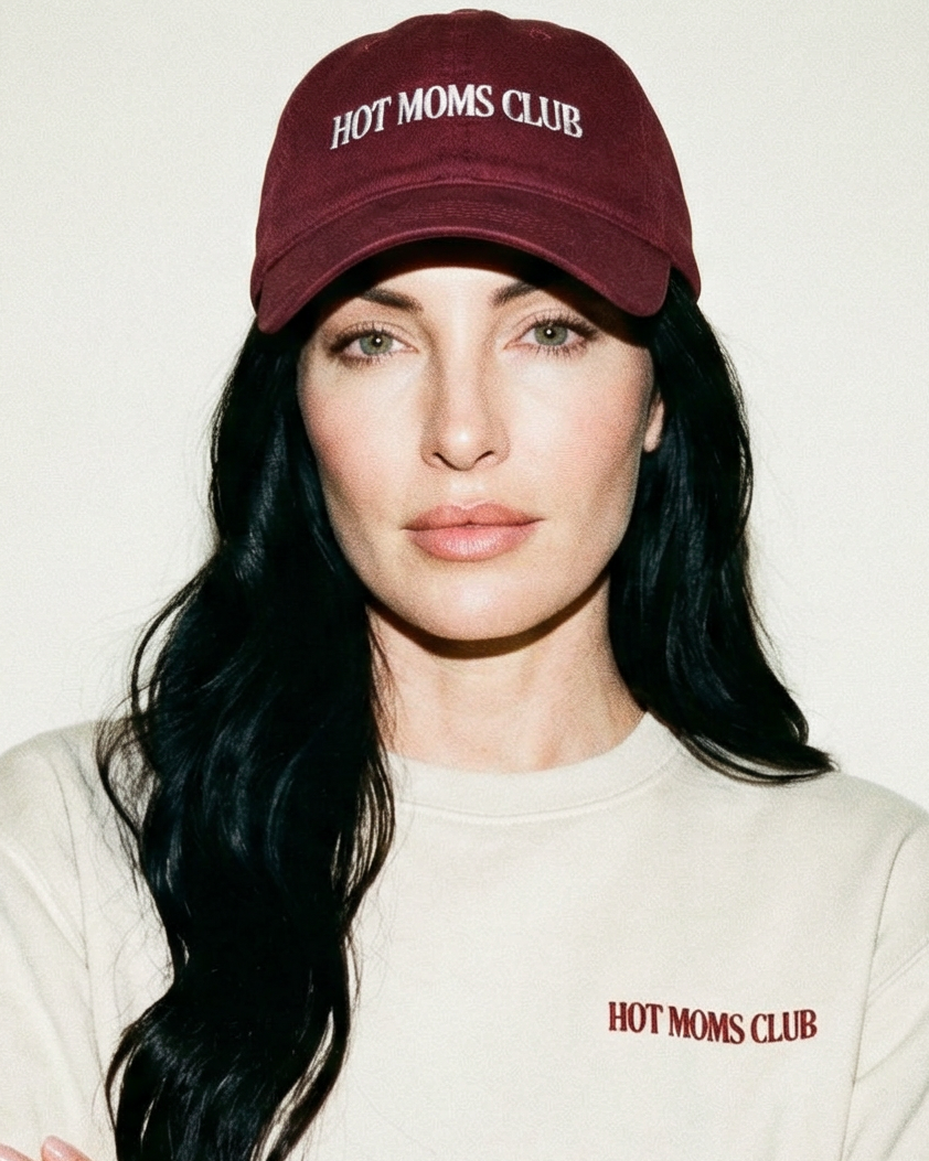 Woman wearing a burgundy 'Hot Moms Club' cap and shirt against a plain background