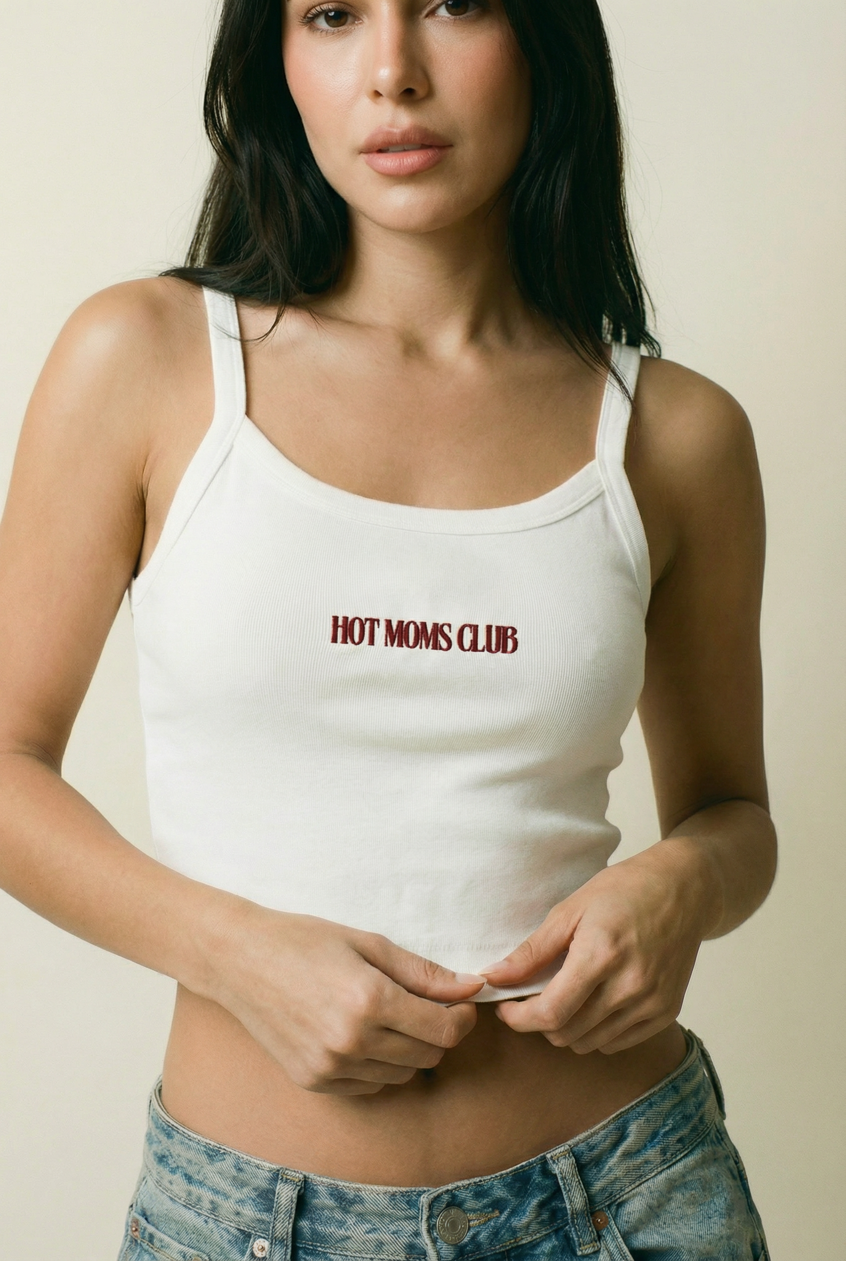 Woman wearing a white tank top with 'HOT MOMS CLUB' text against a plain background