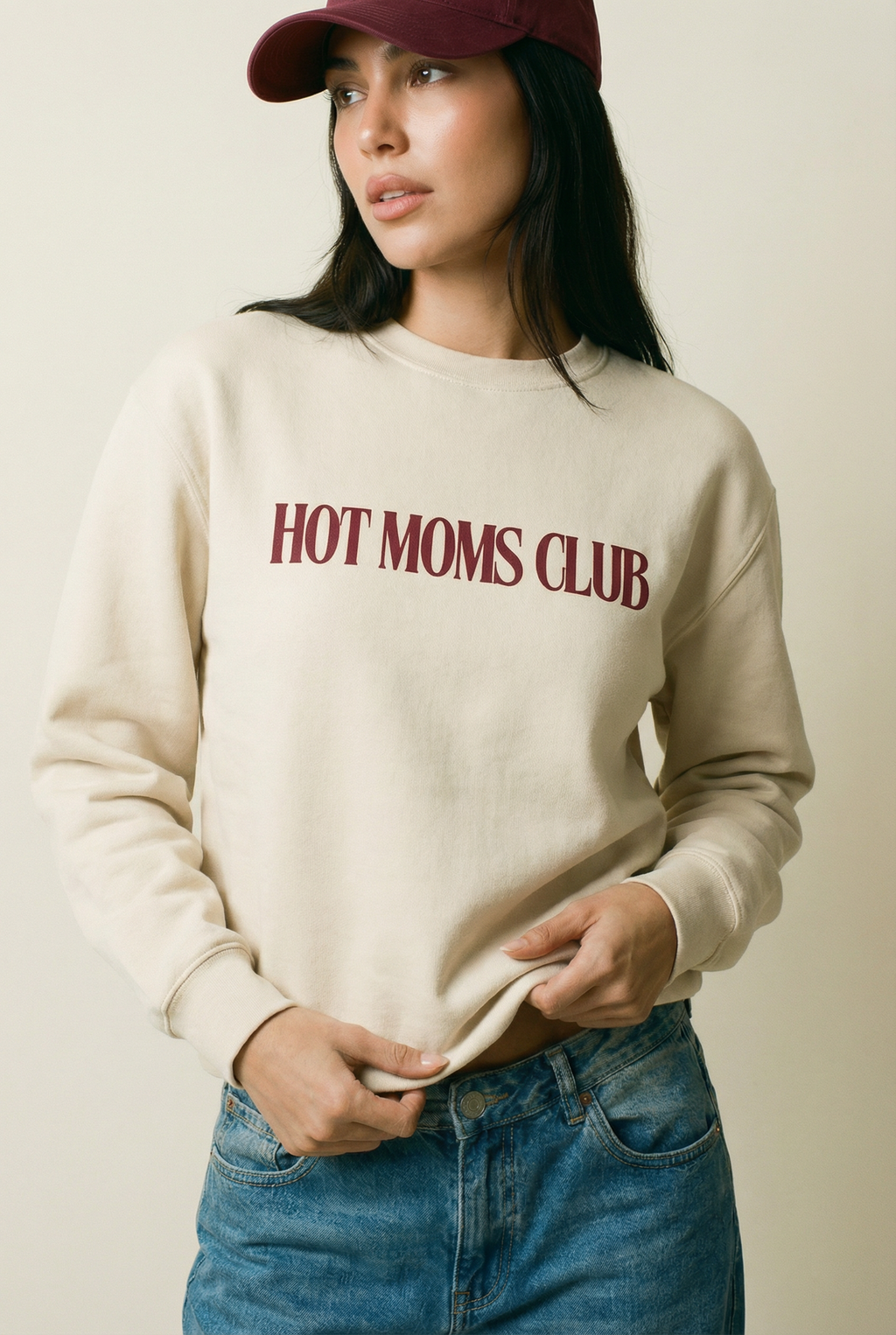 Woman wearing a cream colored crewneck sweatshirt with 'HOT MOMS CLUB' text, blue jeans, and a burgundy cap on a plain background.