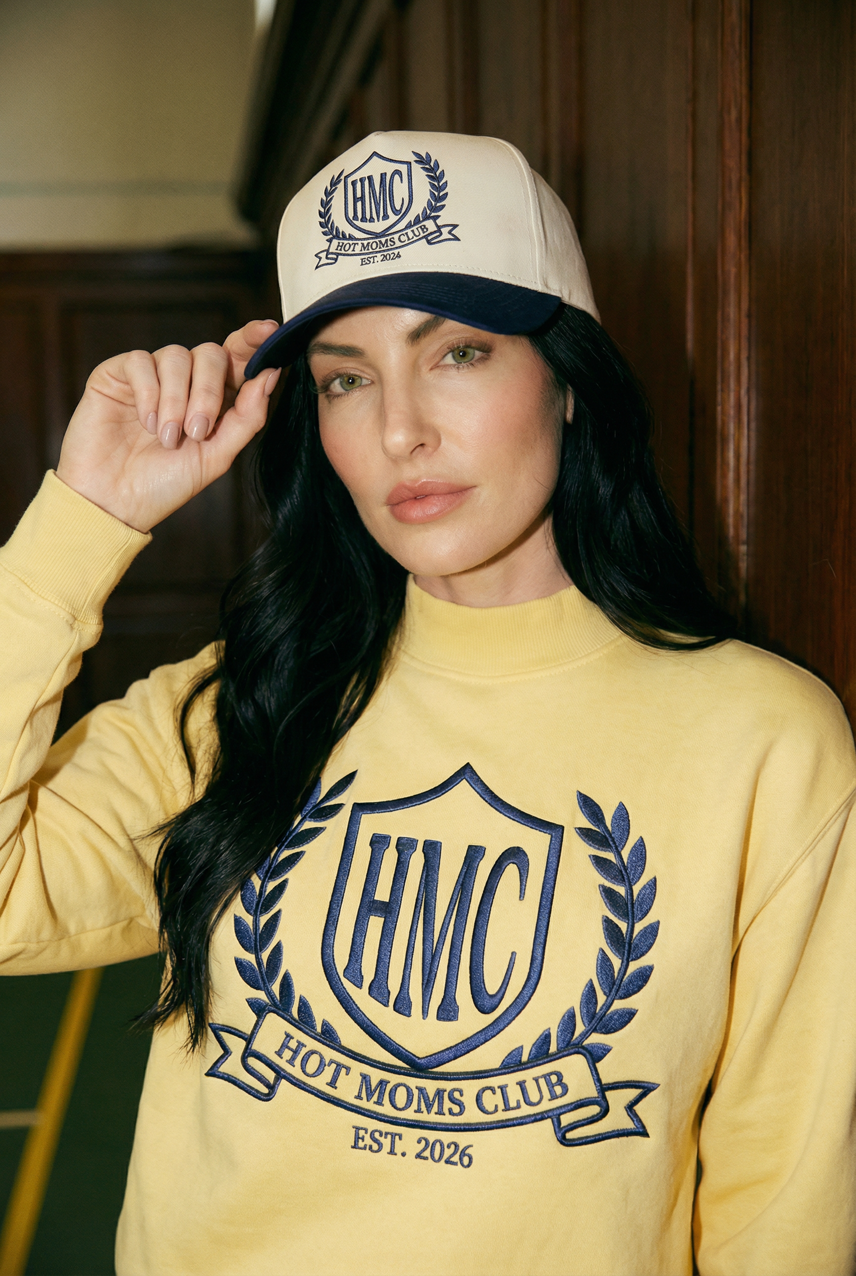 Woman wearing a yellow sweatshirt and cap with 'HMC Hot Moms Club' branding.