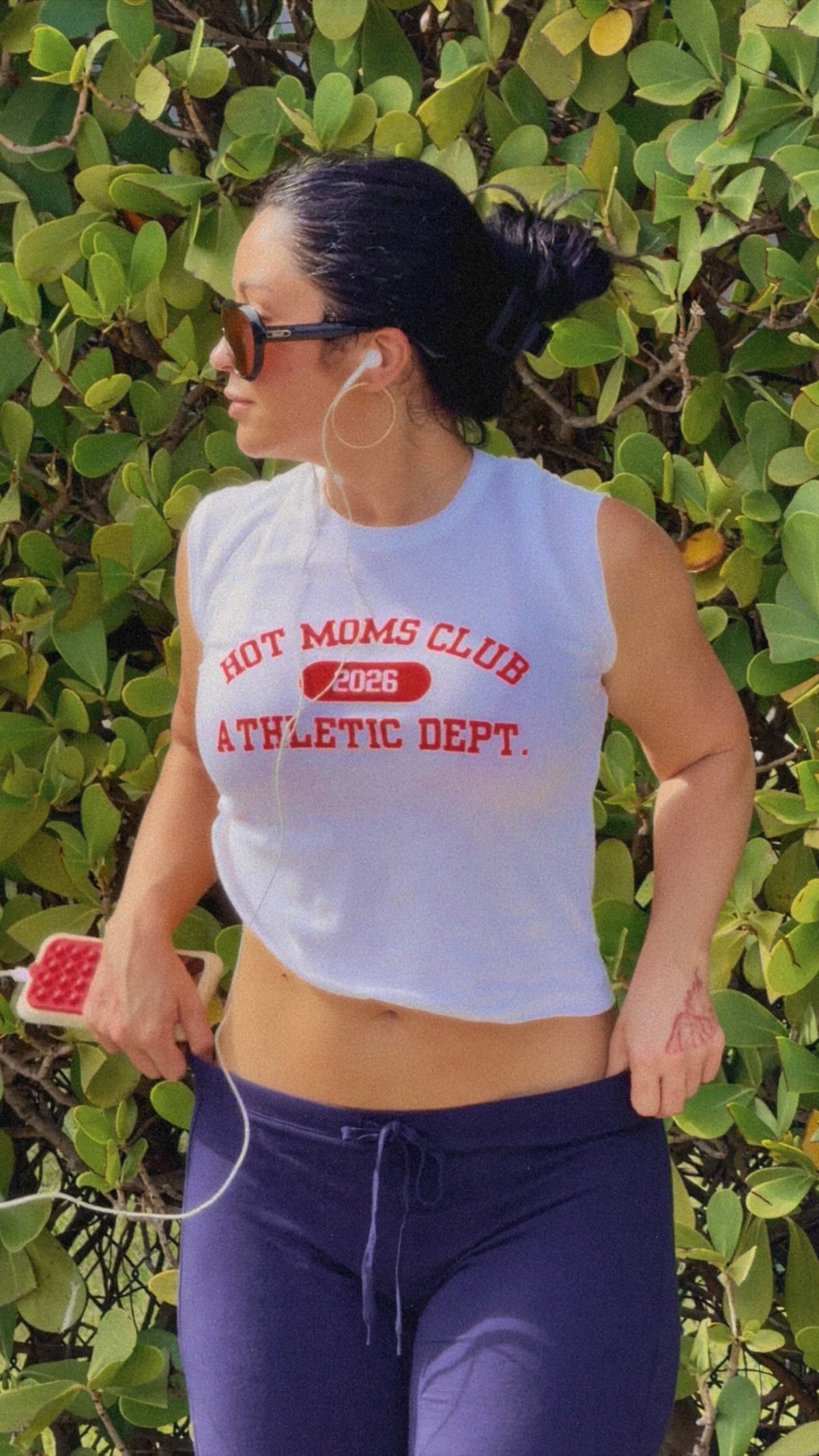A woman wearing a white sleeveless crop top with red text reading 'HOT MOMS CLUB 2026 ATHLETIC DEPT.' and dark purple athletic pants, standing outdoors in front of a green leafy wall.