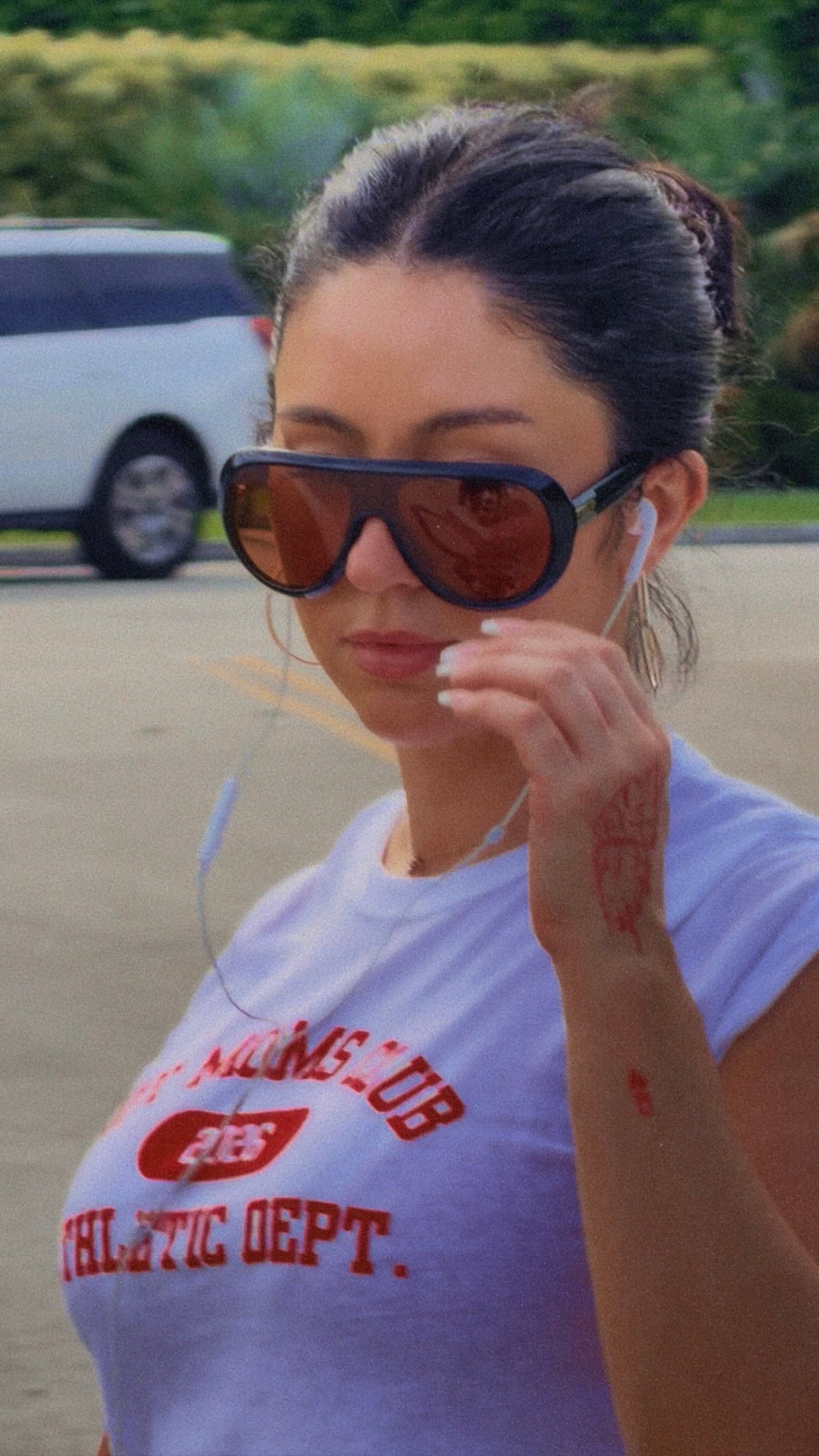A woman outdoors wearing oversized dark sunglasses with orange-tinted lenses and a white graphic T-shirt that reads 'ATHLETIC DEPT.' in red.