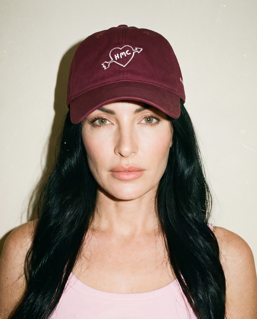 Woman wearing a burgundy baseball cap with HMC logo inside a heart with an arrow through it