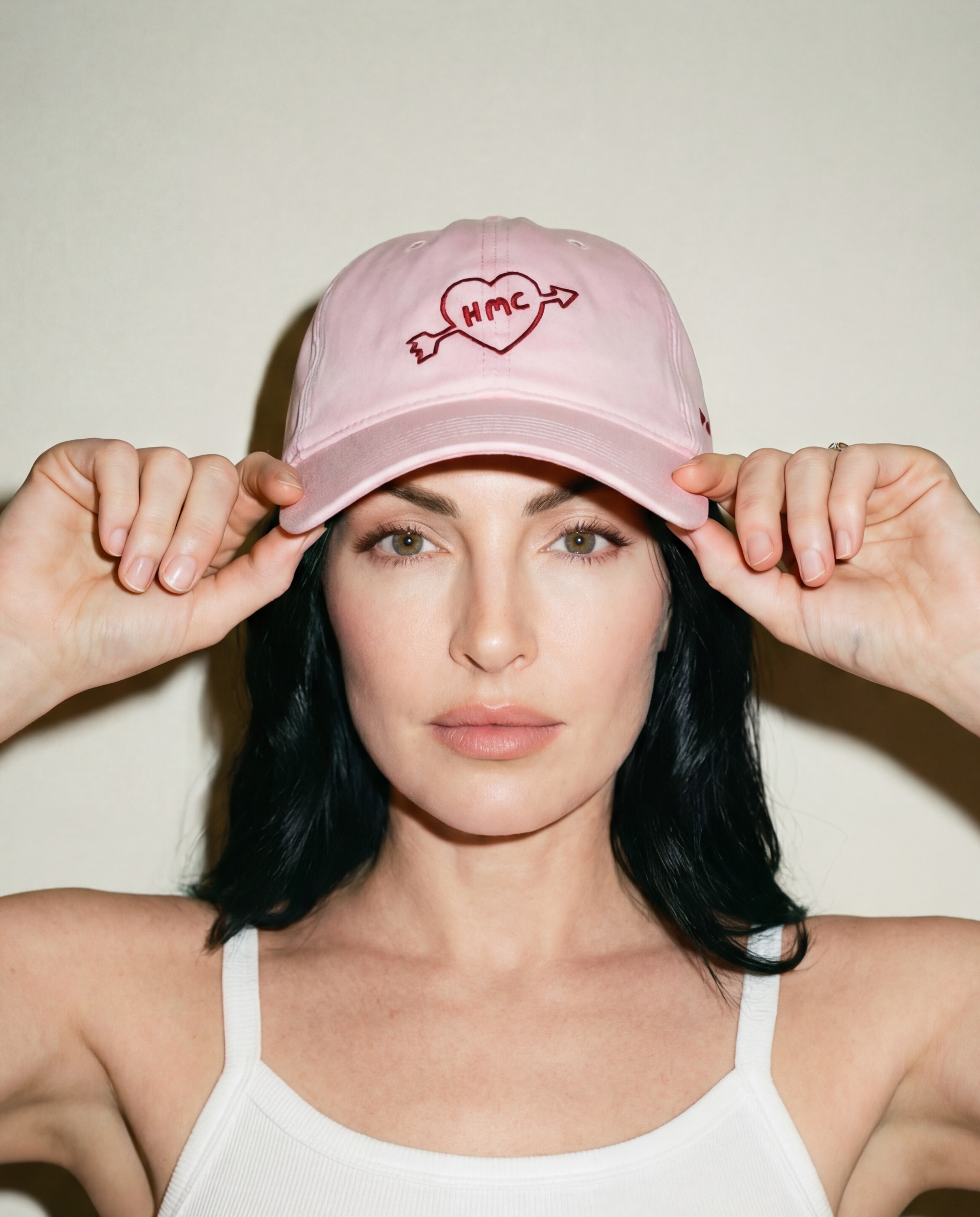 Woman wearing a pink baseball cap with 'HMC' inside a heart with an arrow through it embroidered in burgundy 