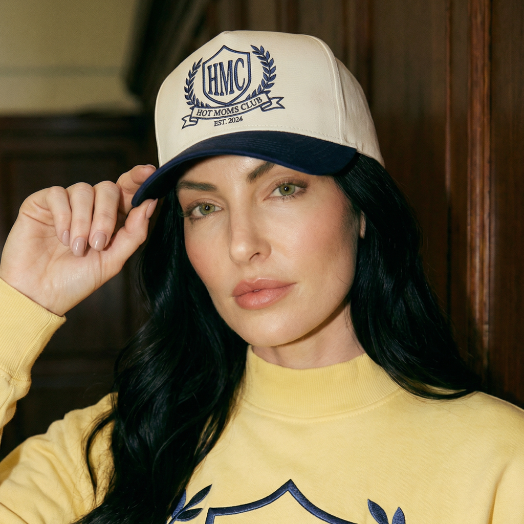 Woman wearing a yellow sweatshirt and cap with 'HMC Hot Moms Club' branding.